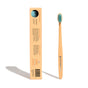 A bamboo toothbrush with turquoise bristles stands upright next to its brown rectangular packaging, which displays product information and a barcode. The package has a circular cutout showing the bristles.