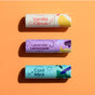 Three lip balm tubes from the Love Your Lips Trio Set by Plant Therapy sit on an orange background. From top to bottom: Vanilla Citrus in white and orange, Lavender Lemonade in purple and yellow, and Cool Mint in light blue and teal, all with playful graphics matching their natural flavors.