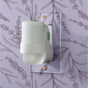 A white Mrs. Meyer's Premium Scented Oil Diffuser Kit is plugged into a wall outlet, set against lavender floral-patterned wallpaper, adding a touch of garden-inspired fragrance to the room.