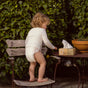 A toddler in a diaper and long-sleeve shirt stands barefoot on a wooden chair outside, reaching for a box of tissues on a small table beside a potted plant, with lush green foliage in the background.