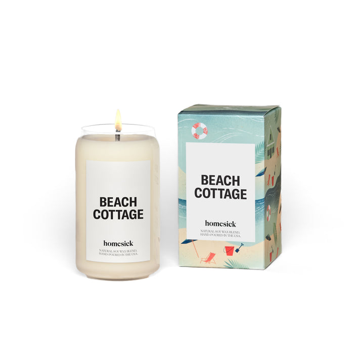 A lit white candle in a glass jar labeled BEACH COTTAGE next to a decorative box with beach-themed illustrations; both display the brand name homesick.