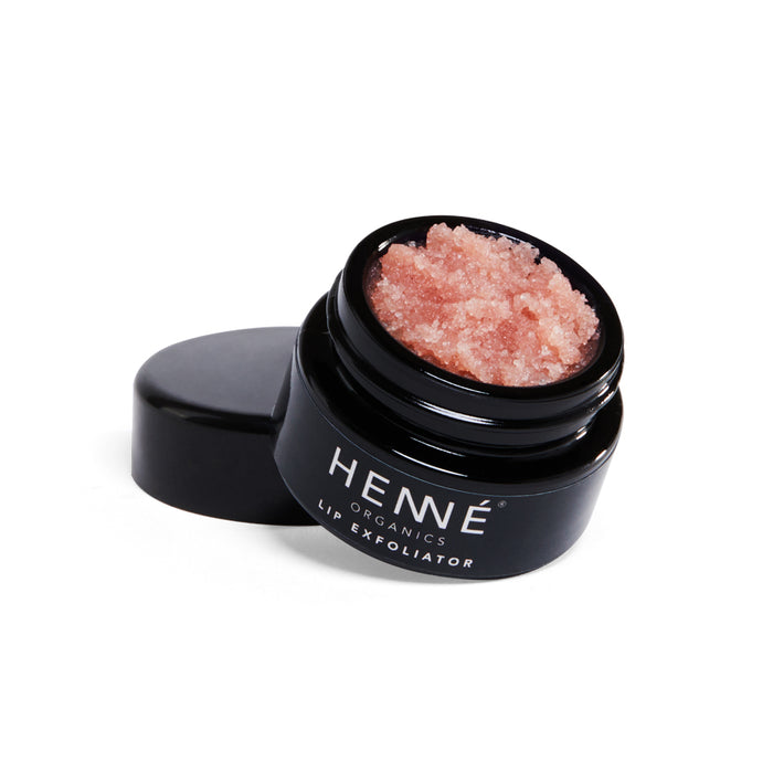 A small black jar labeled Henné Organics Lip Exfoliator is shown with its lid off, revealing a pink, grainy scrub inside. The open lid rests beside the jar on a white background.