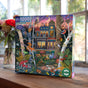 The Alchemist's Forest 1000 Piece Puzzle by eeBoo Piece & Love, featuring Vasilisa Romanenko’s whimsical art of a magical forest with animals, mushrooms, and flowers, is displayed on a wooden windowsill with blooms in the background.