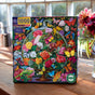The eeBoo Piece & Love Butterfly Still Life 1000 Piece Puzzle box, adorned with vivid butterflies and flowers under a glass dome, sits on a wooden table in a bright room with a vase of fresh flowers nearby.