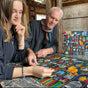 A young woman and an older man sit at a table in a cozy room with bookshelves, working together on the colorful Alchemists Cabinet 1000 Piece Puzzle by eeBoo Piece & Love.