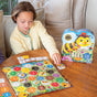A child enjoys playing eeBoo Piece & Love’s Bouncy Bees Shaped Game for ages 3+, which includes vibrant flowers, numbers, and bee tokens. The box features a smiling cartoon bee in a hat. This fun game helps kids learn colors.