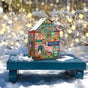 The Christmas House 100 Piece Puzzle by eeBoo Piece & Love features illustrated rooms and characters. Its colorful house-shaped box shines on a blue bench in snowy outdoors with glowing lights, perfect for a holiday family activity.