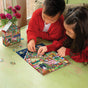 Two children sit at a green table working on the eeBoo Piece & Love Christmas House 100 Piece Puzzle. The house-shaped box and a vase of red flowers are also on the table.