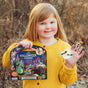 A red-haired girl in a yellow sweater smiles outdoors, holding an eeBoo Piece & Love Kids Sticker Book and Halloween Stickers. Some of the Halloween Stickers shimmer with holographic foil for extra festive fun.