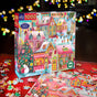 The eeBoo Piece & Love Holiday Village 1000 Piece Puzzle, made from sustainably sourced materials, features a festive village scene. The colorful box sits on a table with scattered pieces and blurred twinkling lights in the background.