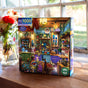 The eeBoo Piece & Love Midnight Kitchen 1000 Piece Puzzle, featuring a colorful kitchen scene, sits on a wooden windowsill beside flowers with sunlight streaming through the window behind it.
