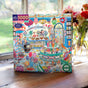 The eeBoo Piece & Love Sweet Shoppe 1000 Piece Puzzle, made from recycled board, features Emily Sutton’s colorful illustration of a candy shop on a sunlit windowsill with blurred flowers in the background.