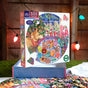 The eeBoo Piece & Love Christmas Cookies 500 Piece Puzzle, featuring Hazel Jarvis’s festive artwork, sits on a table with holiday lights and decorations glowing in the background.