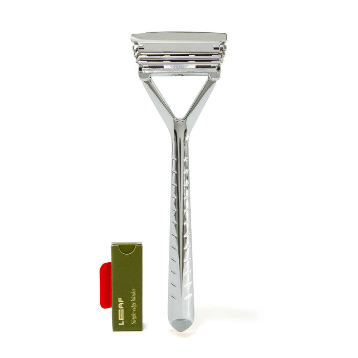 A sleek silver metal razor with three blades is standing upright next to a small green and red box labeled LEAF. The background is white.