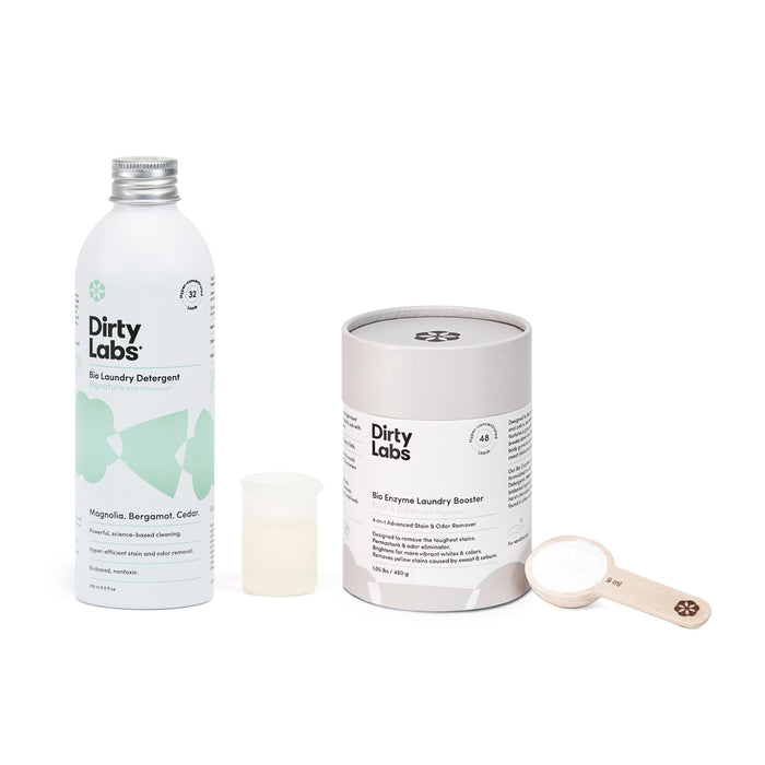 A bottle of Dirty Labs Bio Laundry Detergent with Phytolase® sits beside a cylindrical Bio Enzyme Laundry Booster, featuring an open lid and wooden scoop. This enzyme-powered duo offers a minimalist, eco-friendly design ideal for sustainable cleaning.