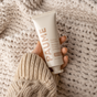 A hand in a cozy beige knit sweater holds a PAUME Probiotic Hand Balm tube with natural probiotics against a textured cream-colored knit backdrop. The minimalist design features beige text, emphasizing its nourishing essential oil blend.