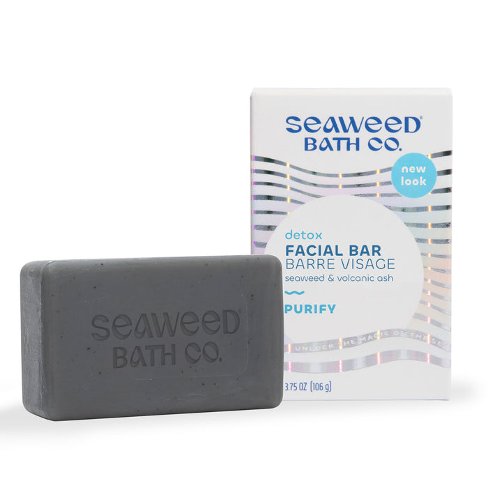 A rectangular gray bar of soap embossed with Seaweed Bath Co. is shown next to its white and iridescent box labeled detox facial bar with seaweed and volcanic ash.