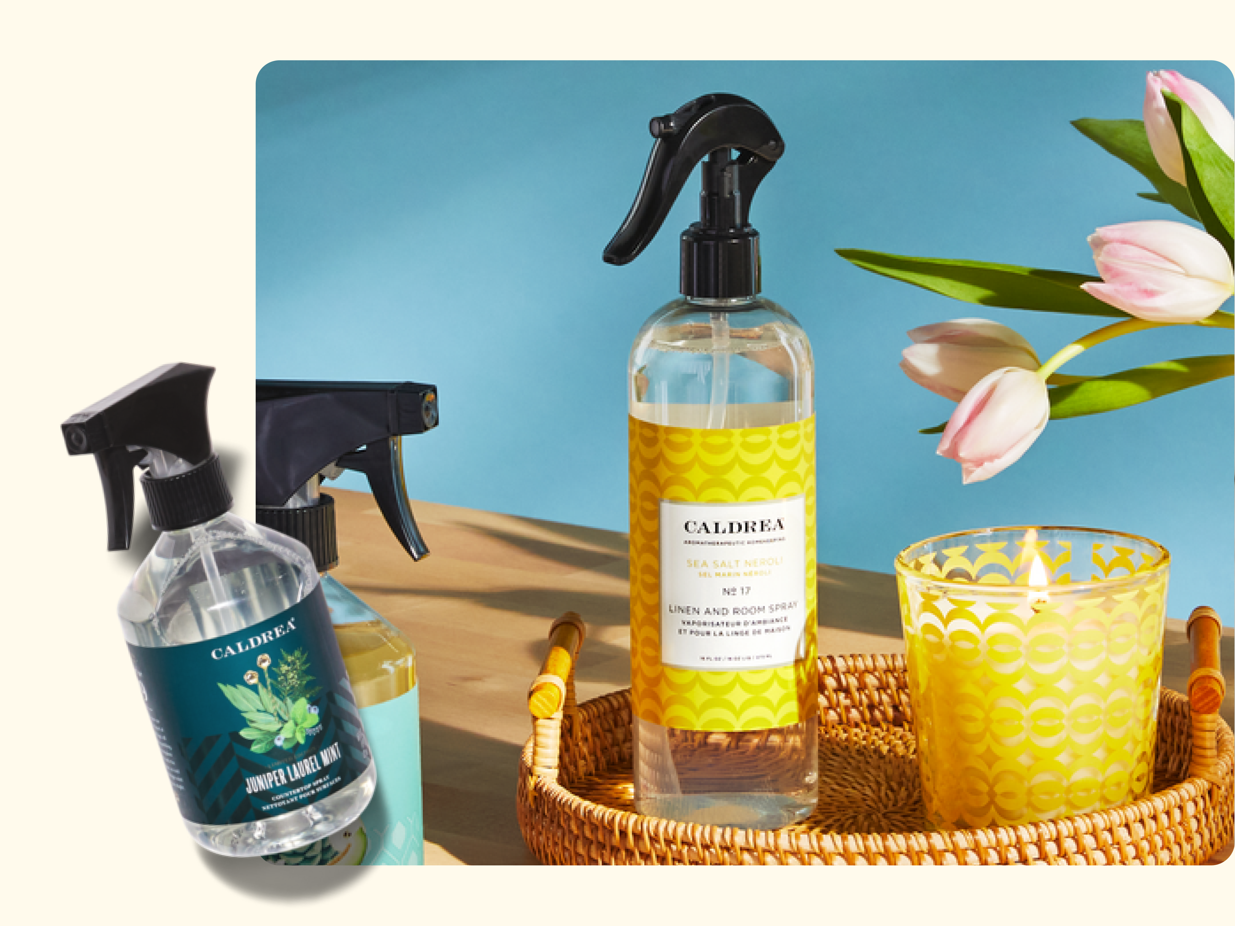 Two spray bottles of Caldrea linen and room spray are shown. One is upright in a wicker tray with a yellow patterned candle and pink tulips, while the other lies at an angle on a off-white surface. Blue background.