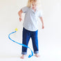 A child with long blond hair, wearing a light blue shirt and rolled-up jeans, joyfully engages in open-ended play. Barefoot against a white backdrop, they twirl the Star Streamer by Sarahs Silks—a blue ribbon wand with a yellow star—enhancing gross motor skills while creating colorful patterns.