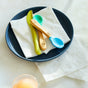 A blue plate holds two folded Grove Co. Tree-Free Napkins made from 100% bamboo, a yellow knife, and two small spoons—one wooden and one blue. Sunlight illuminates the surface where extra sustainable bamboo napkins and a glass of orange juice sit nearby.