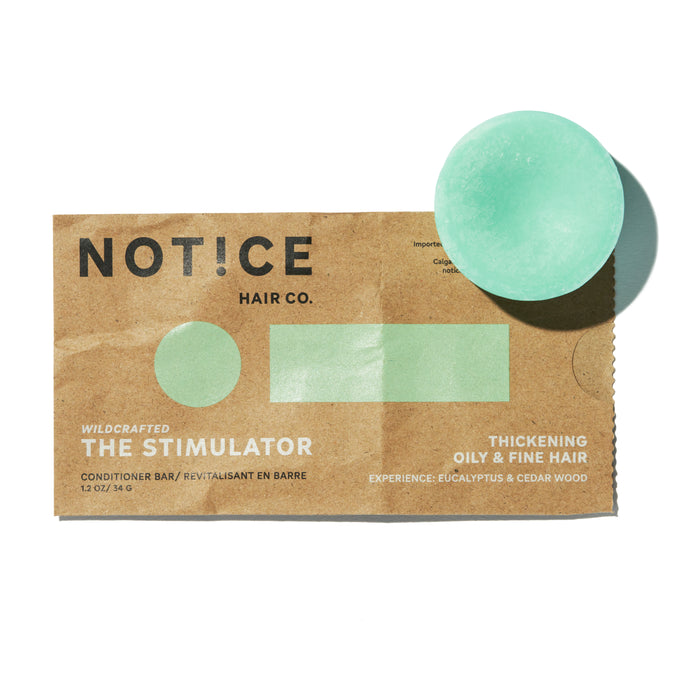 A round, light green conditioner bar sits beside its brown paper packaging labeled NOTICE Hair Co. THE STIMULATOR, for thickening oily & fine hair, with eucalyptus and cedar wood.