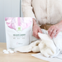 A person in a beige shirt handles a white knitted fabric near Grab Greens Delicate Laundry Detergent Pods. The package, with its natural ingredients and white and pink accents, highlights green chemistry as several pods sit on the wooden surface.