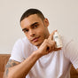 A man in a white t-shirt sits on a couch, smiling slightly and holding up a small white bottle labeled Phil's Daily Facial Cleanser. He points at the bottle with one finger.