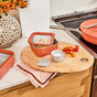 A kitchen scene with a coral-colored food container holding salad, a small bowl of salt, sliced cherry tomatoes, a pink napkin, a large vase with orange flowers, and a matching coral pan on the stove.