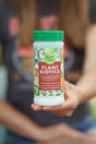 A person holds a container labeled Good Dirt Plant Biotics with a green lid and colorful floral design, intended for indoor and outdoor plant use. The background is blurred.