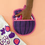 A hand reaches into The Neighborgoods Pot Holder - Fig, shaped like a fig, on a peach background. Next to it is a white towel with vibrant berry designs and “JIGGY” printed—both playful kitchen essentials.