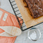 Brownies cool on Full Circles Compostable Parchment Paper Roll atop a black wire rack. Two wooden spoons, one with engraved text, sit on salmon fabric. A small sieve with powdered sugar lies nearby on marble.