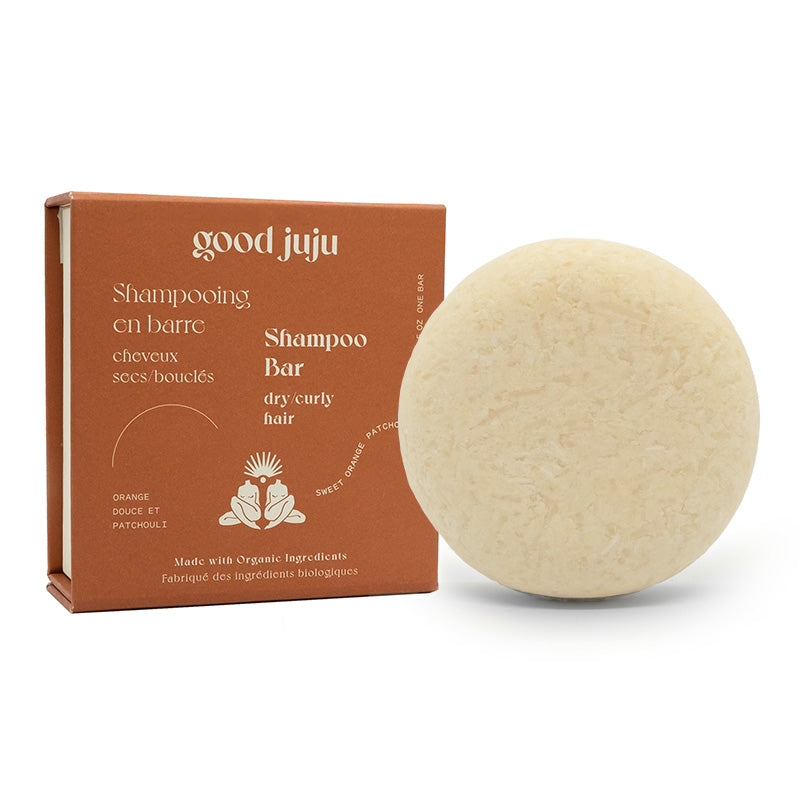 A round Dry/Curly Shampoo Bar lies beside its brown box packaging, labeled Good Juju Body & Home. Made with organic ingredients for frizz control, the bilingual package features illustrations of orange, patchouli, and hair with text in English and French.