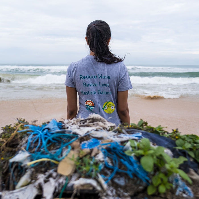 A person with long dark hair stands on a beach facing the ocean, wearing a gray shirt that reads Reduce Waste, Revive Lives, Restore Balance. Piles of marine debris and ropes are scattered on the sand in front of them.