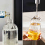 Split image: Left side shows a bottle of Grove Co. liquid being poured into a clear pump dispenser. Right side shows the dispenser being filled with water from a faucet; the dispenser has a yellow base labeled GLASS.