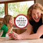 A laughing woman and child play with flour at a kitchen counter as sunlight streams in. A round sign reads, “Life is messy. Get your hands dirty.” Nearby, Mrs. Meyer’s Dish Soap stands ready for quick cleanup.