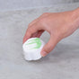 A hand holding a round white cleaning tablet with a green stripe, positioned above a light gray tiled surface.