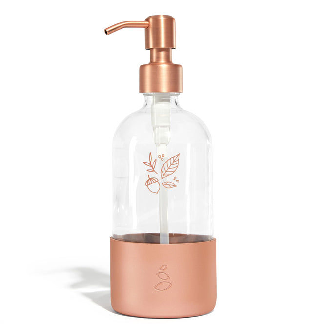 The Grove Co. Refillable Dish Soap Dispenser features a transparent body with a bronze pump and base, adorned with a leaf and fruit design. It adds elegance to your sustainable home, reducing plastic waste while sitting beautifully against any plain white backdrop.