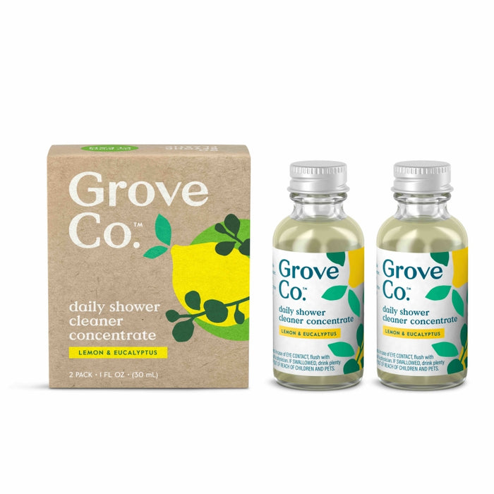 Two glass bottles of Grove Co. daily shower cleaner concentrate (lemon & eucalyptus scent) are next to a brown cardboard box with green and yellow leaf designs and product information.