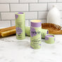 A lavender lotion bar in a light green and purple tube labeled erleia stands upright on a bathroom counter, with another tube, a wooden hairbrush, a nail brush, and a white vase in the background.