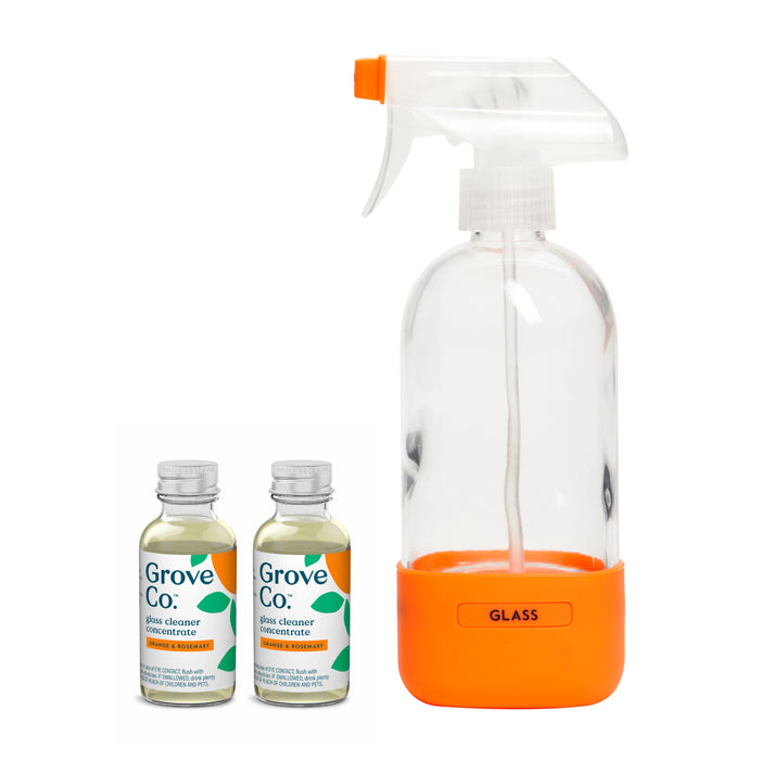 A glass-cleaner-spray-bottle with a labeled orange base is beside two small Grove Collaborative glass cleaner concentrate bottles, featuring white caps and green text.