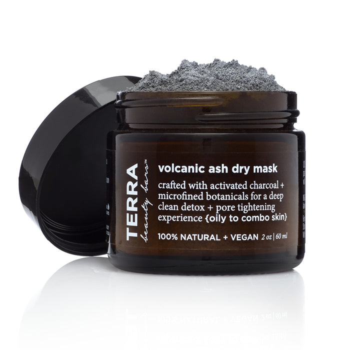 A brown jar labeled Terra Volcanic Ash Dry Mask sits open with gray powder overflowing, showing product texture. The label highlights it as natural, vegan, and suitable for oily to combination skin.
