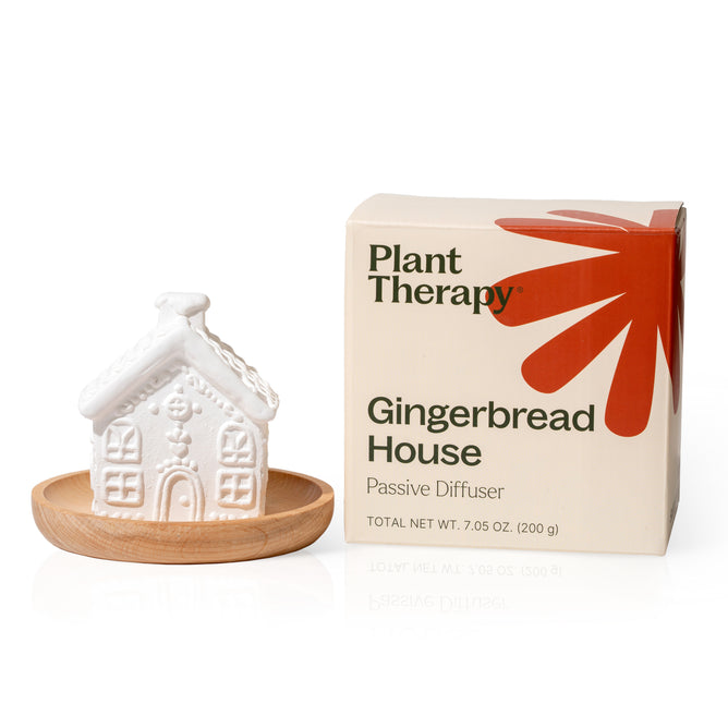 A white ceramic Gingerbread House Passive Diffuser by Plant Therapy rests on a wooden tray beside its cream and orange box with an abstract design—an ideal choice for festive holiday décor.