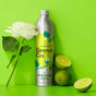 A metal bottle of Grove Co. dish soap with citron & white rose scent stands next to a white rose and two limes, one whole and one halved, against a bright green background.