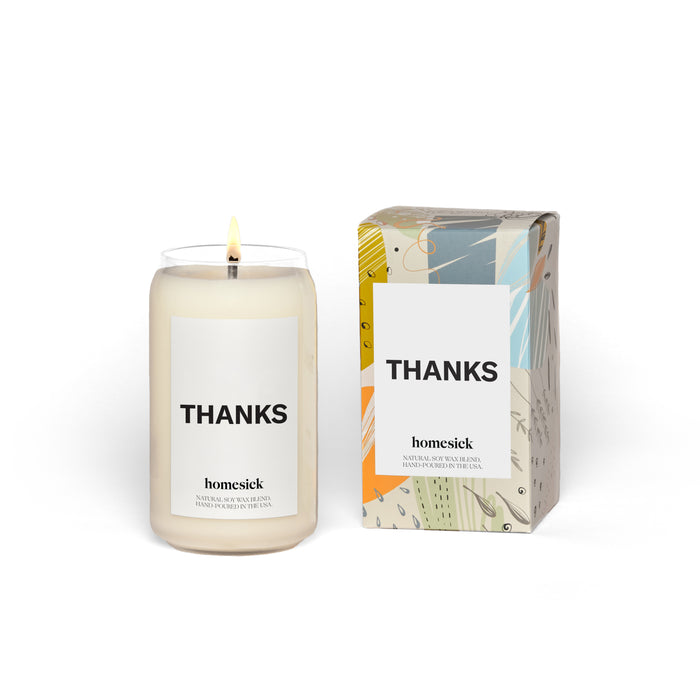 A lit white candle in a glass jar labeled “THANKS” stands next to its colorful, patterned box, which also displays “THANKS” and “homesick” branding.