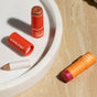 Three Axiology makeup products on a beige marble surface: two orange cylindrical sticks with exposed makeup (one pink, one brown) and one small crayon-shaped pencil, with part of a white dish visible.