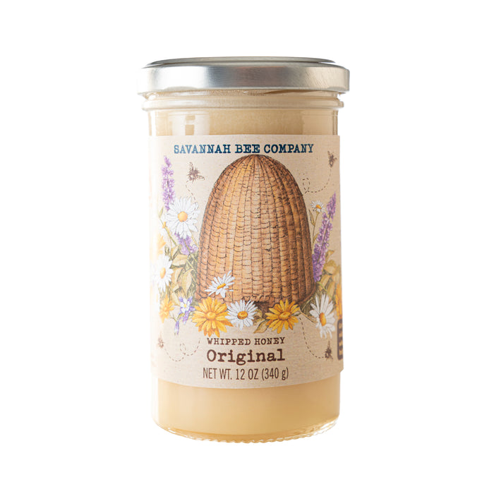 Savannah Bee Companys Whipped Honey Original comes in a 12 oz (340 g) clear glass jar, ideal for baked goods. The label displays a skep with illustrated flowers and bees, and the creamy honey inside is light in color.