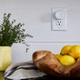 A white smart plug is plugged into a wall outlet above a counter with a yellow potted herb, a striped cloth, and a wooden bowl filled with lemons.
