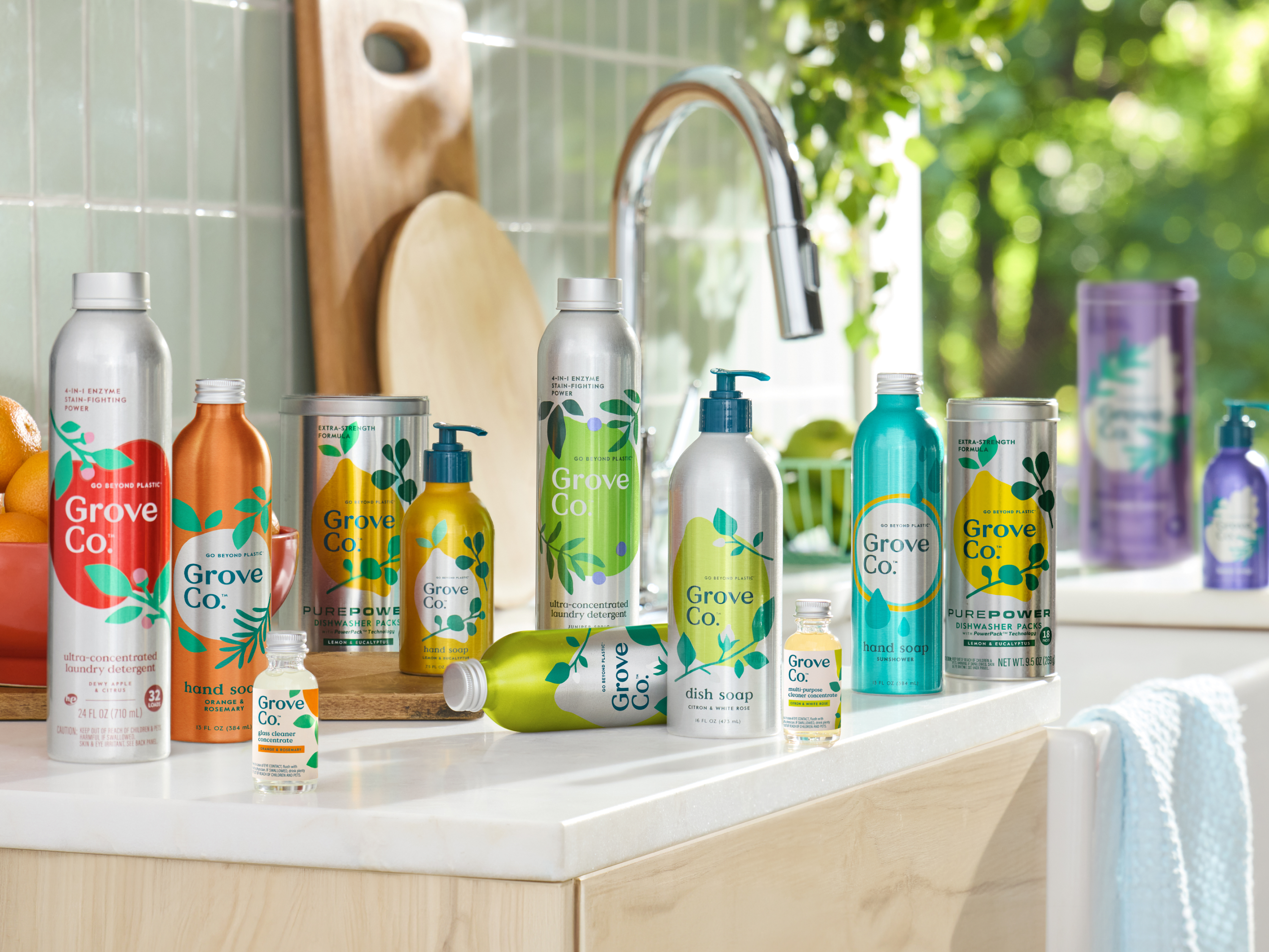 Group of Grove Co. products on a kitchen counter. Group includes aluminum and glass solutions – aluminum laundry detergent, dishwasher pods, hand and dish soap, and glass cleaner concentrates. Products are arranged in color order of the rainbow. Light floods from a window behind.