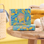Two boxes of Grove Co. Laundry Sheets - Spring Collection, with a blue and yellow floral design, rest on a wooden table next to yellow towels, a wicker basket with towels, and cotton swabs, all against a vibrant yellow wall.