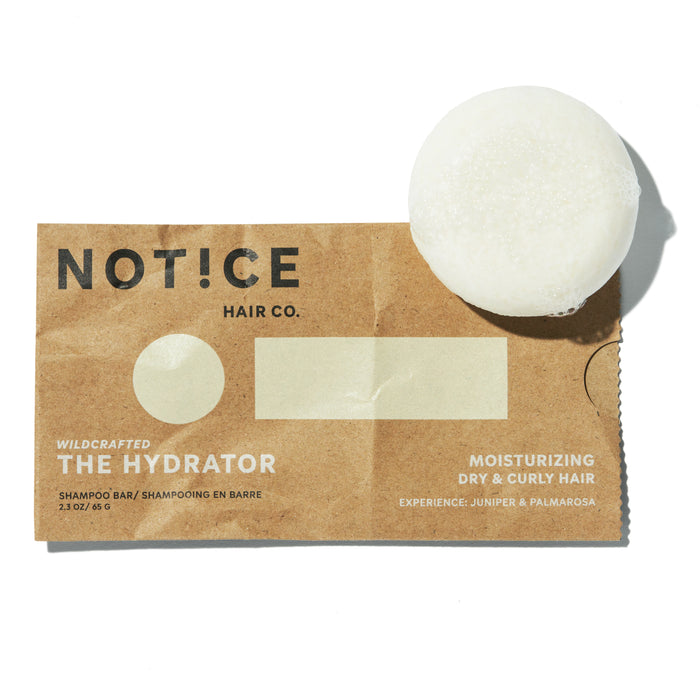 A round white shampoo bar rests on a brown paper packaging labeled NOT!CE Hair Co. The Hydrator for moisturizing dry and curly hair, with juniper and palmarosa scent.